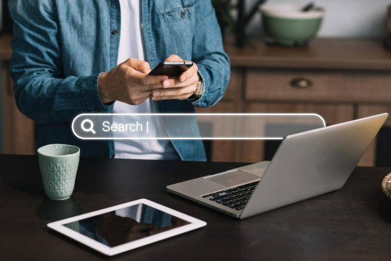 Persona utilizando un celular frente a una computadora portátil, con una barra de búsqueda digital interactiva superpuesta en primer plano. El SEO y visibilidad en IA ya forman parte de una misma estrategia de posicionamiento digital.